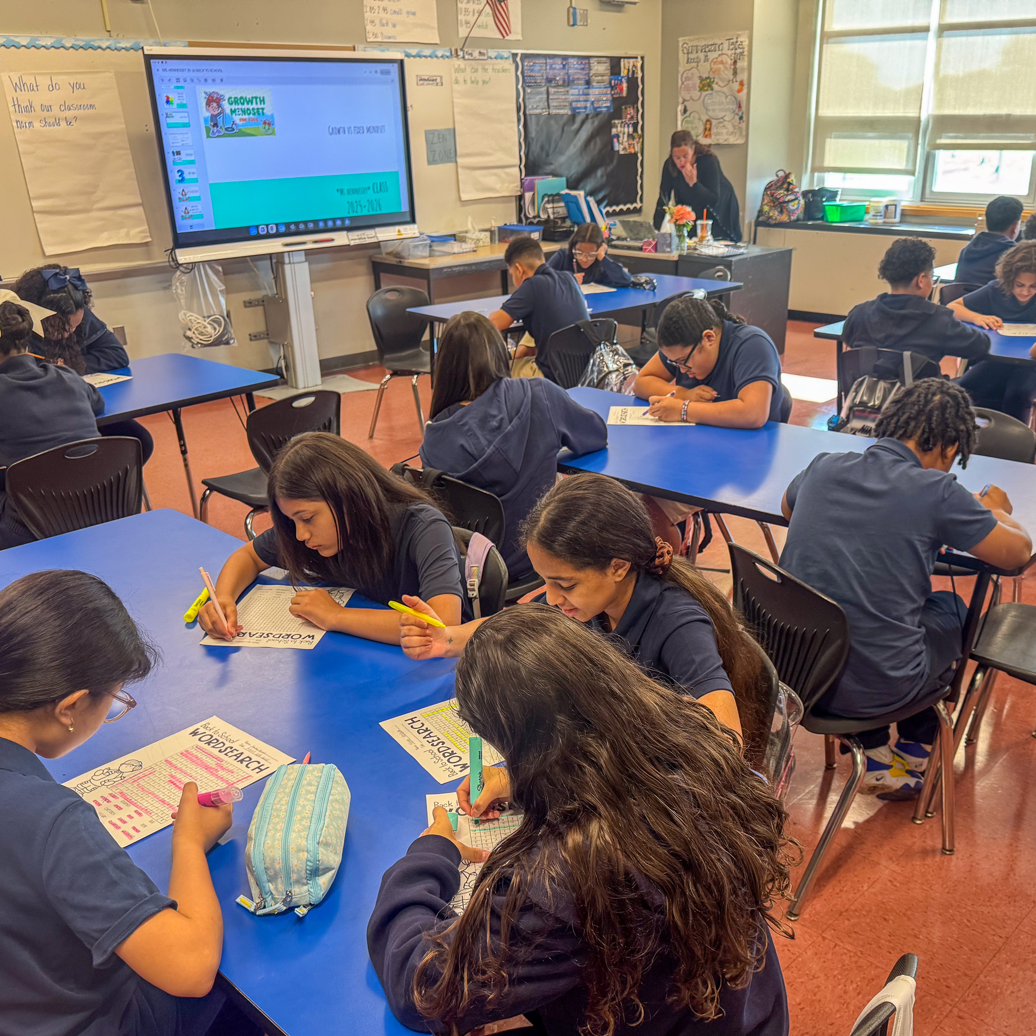 Middle school students in navy uniforms sit at blue tables, writing on worksheets with highlighters and pencils.