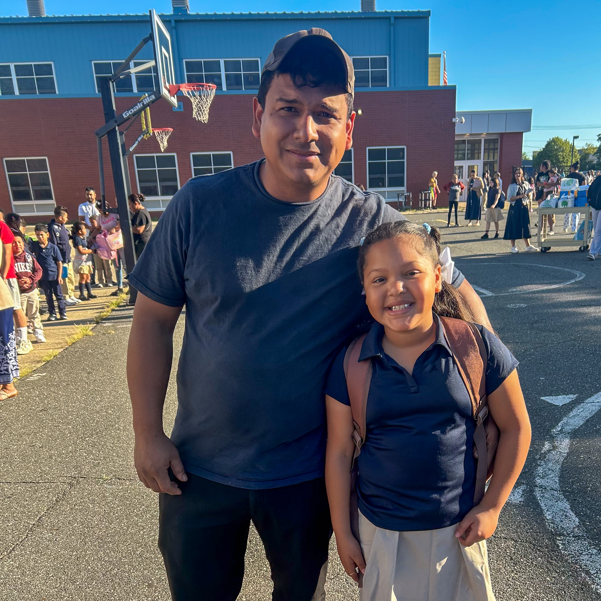 A smiling student in a navy polo and khaki skirt stands with her parent in front of the school building on the first day of school.